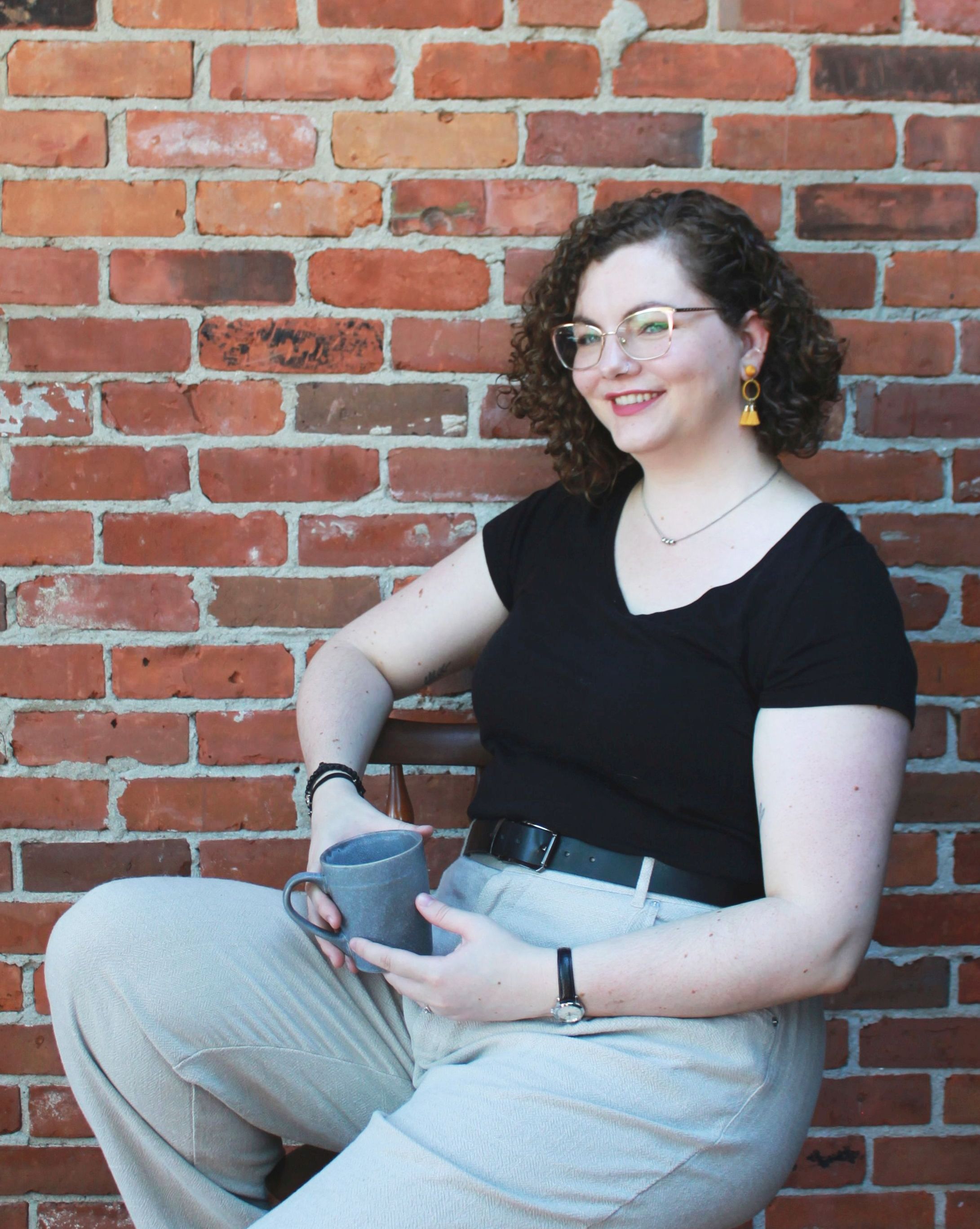 Rebecca McIntyre leans back in a chair, holding a blue mug. They are smiling wide and looking to one side. They are wearing a black t-shirt and beige trousers.