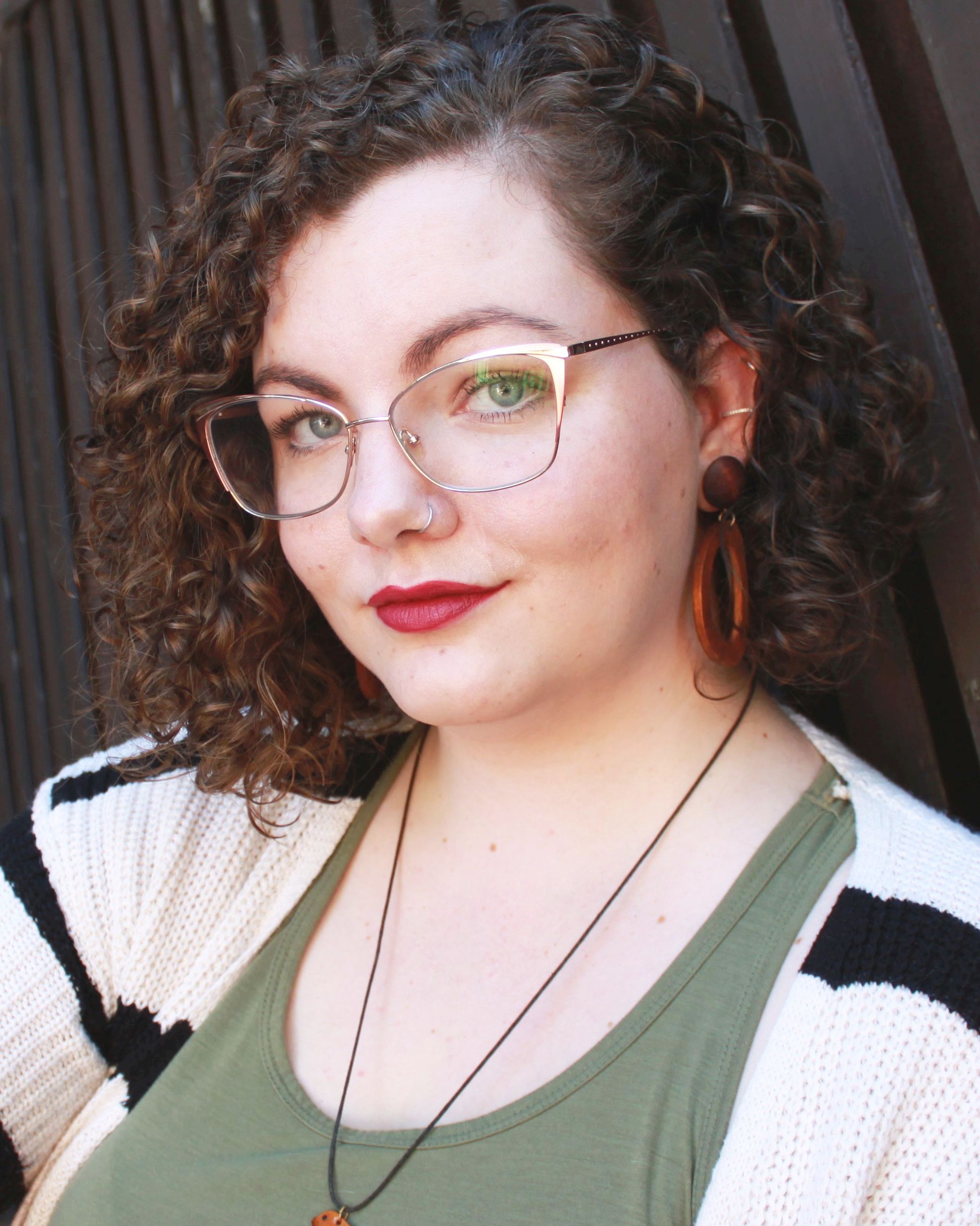 A headshot of Rebecca McIntyre, smiling gently. Their shoulder-length, curly brown hair is pulled back on one side. They are wearing a green shirt and a black-and-white cardigan.