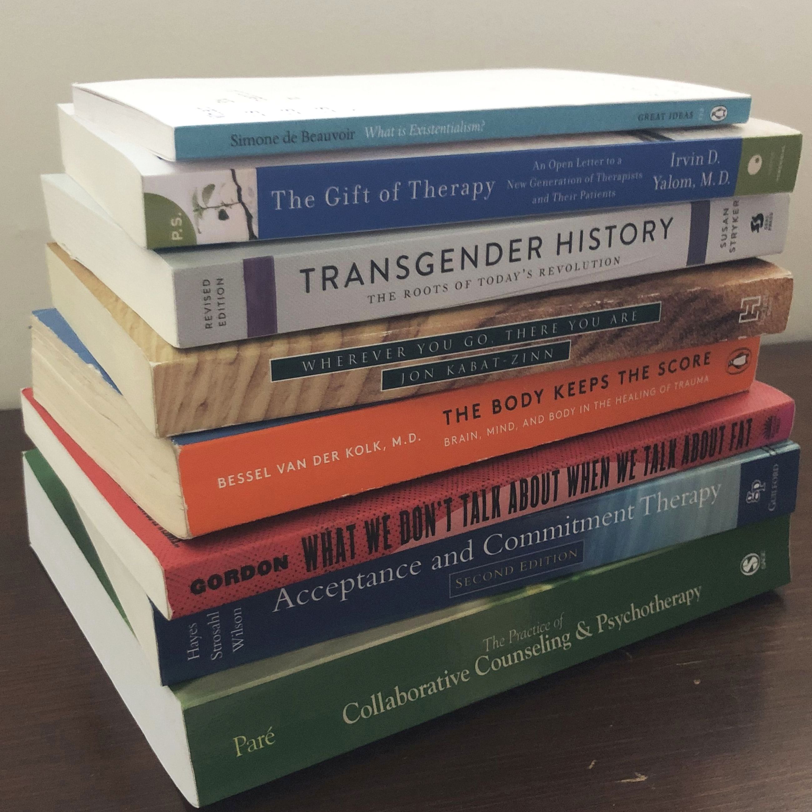 A stack of books. The following titles and authors are visible: ‘What is Existentialism?’ by Simone de Beauvoir; ‘The Gift of Therapy’ by Irvin D. Yalom; ‘Transgender History’ by Susan Stryker; ‘Wherever You Go, There You Are’ by Jon Kabat-Zinn; ‘The Body Keeps the Score’ by Bessel van der Kolk; ‘What We Don’t Talk About When We Talk About Fat’ by Gordon; ‘Acceptance and Commitment Therapy’ by Hayes, Stroshal, and Wilson; and ‘Collaborative Counseling and Psychotherapy’ by Paré.